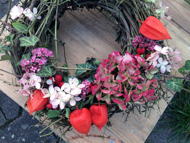 Selbst gebundener Herbstkranz mit rosa Blüten, orangeroten Lampions und grünen Blättern auf einem Holzbrett